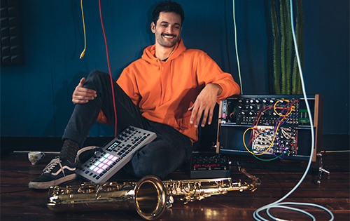 Dillon sitting relaxed with electronic music equipment and a saxophone, showcasing his unique style