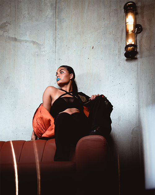 Model in black with striking blue lipstick, lounging thoughtfully with an orange jacket