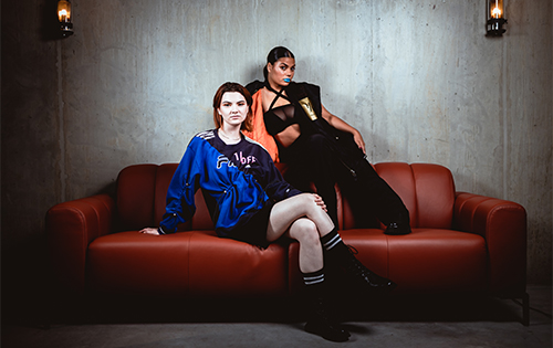 Two fashion models posing with attitude on a red leather couch in sporty and stylish outfits