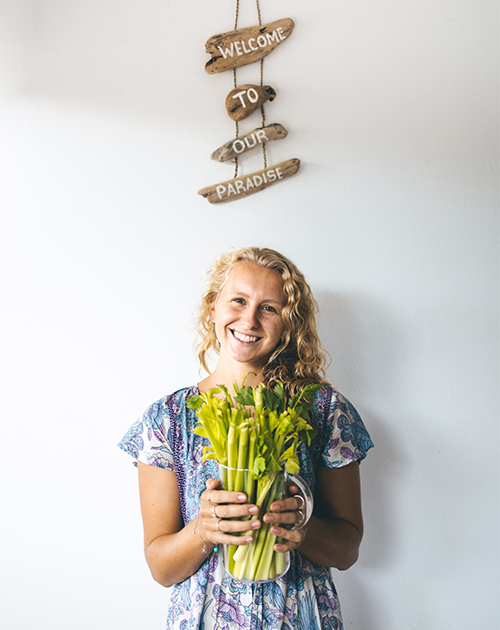 Chef Maddie holding a fresh bunch of green stems with a welcoming smile, behind a 'Welcome to Our Paradise' sign at a Fuerteventura retreat