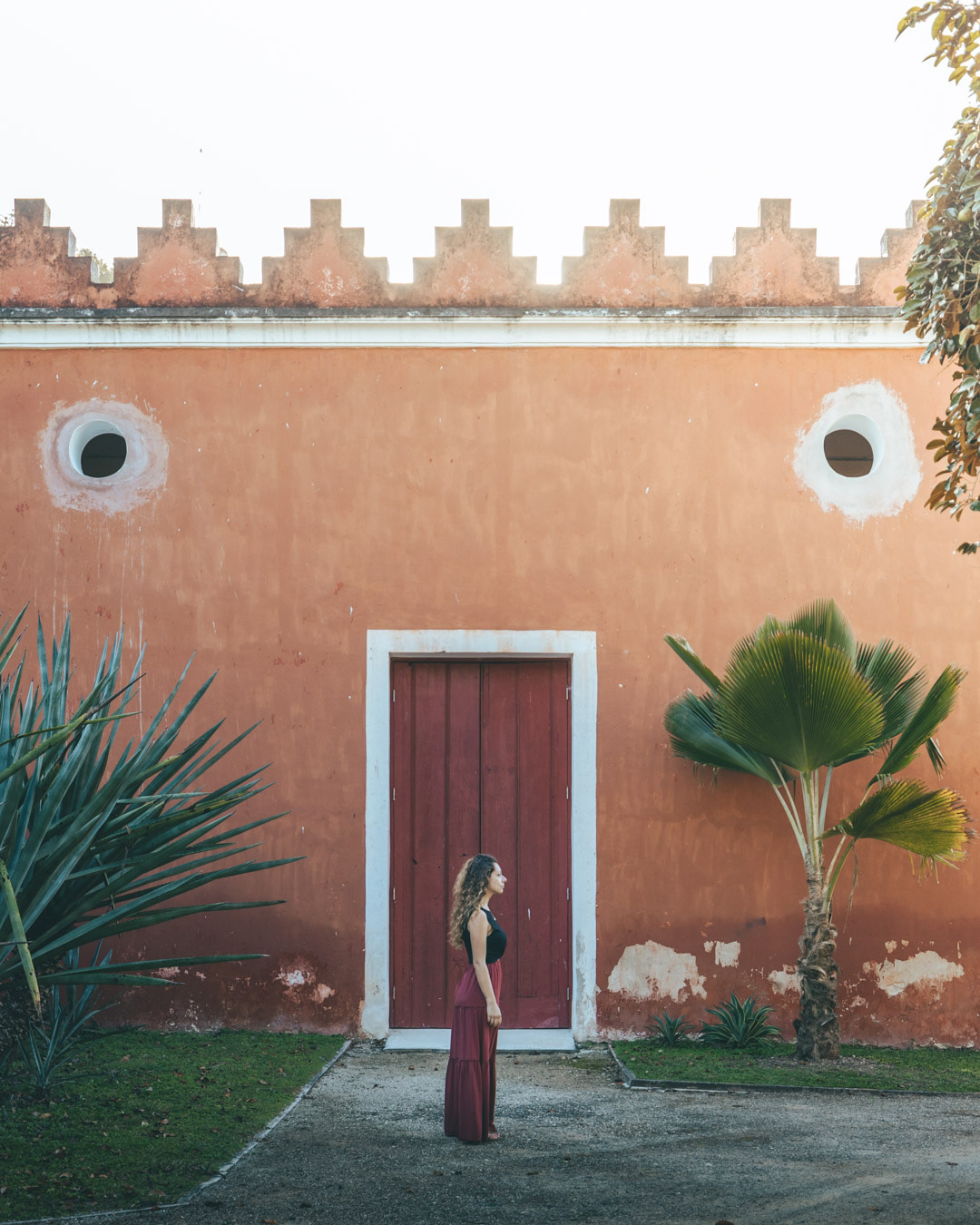 Woman in red pondering by the rustic red wall with palm tree at Hacienda San Antonio Millet