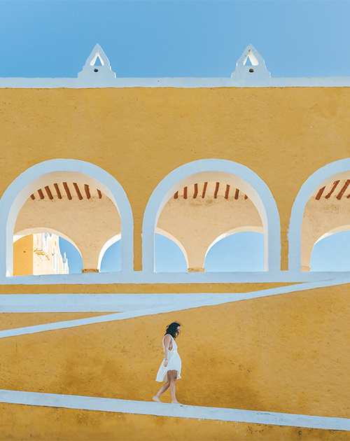 A woman in white dress walking through the vibrant yellow and white arches of the colonial architecture in Yucatan, Mexico