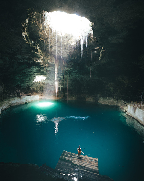 Tranquil cenote waters illuminated by a natural light beam, with a visitor on a wooden platform in Yucatan's natural sinkhole