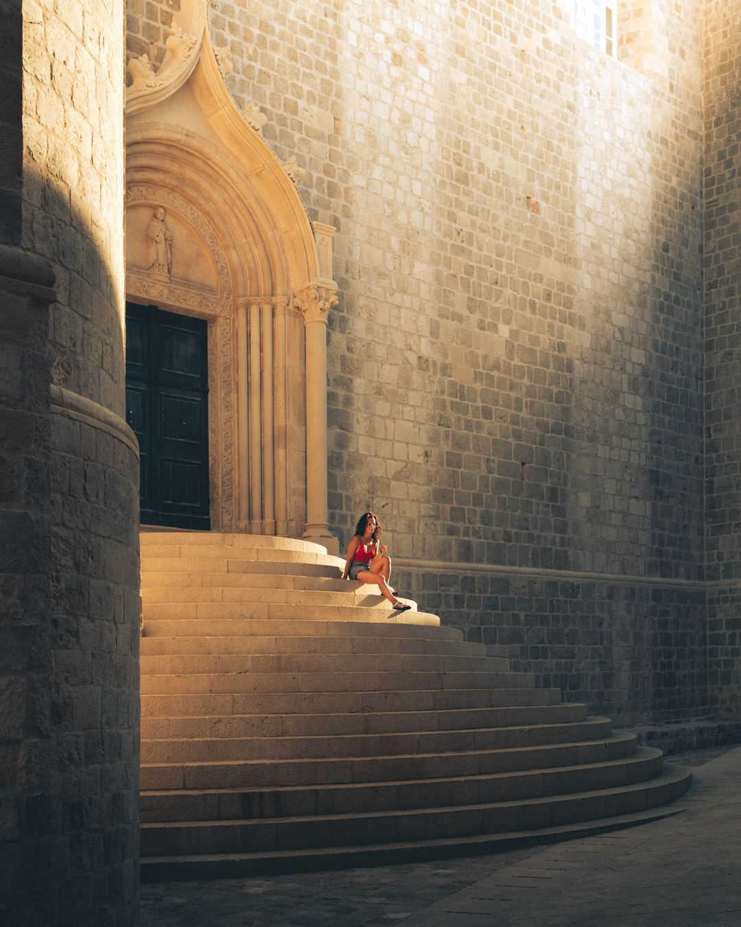 A thoughtful portrait of a person sitting on the ancient steps of a Dubrovnik church, bathed in sunlight