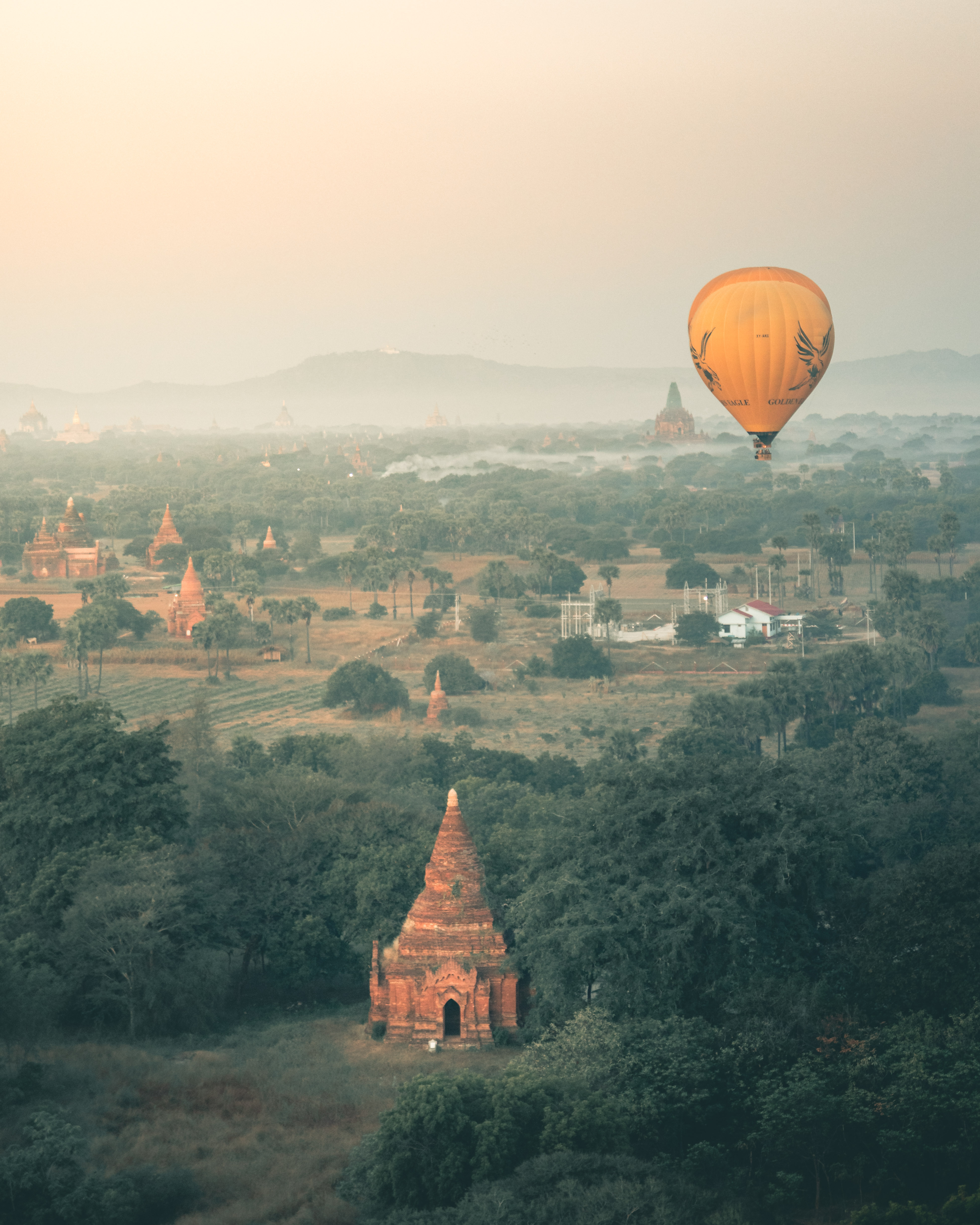 A majestic hot air balloon rising above the misty dawn landscape of Myanmar, dotted with ancient temples and lush greenery.